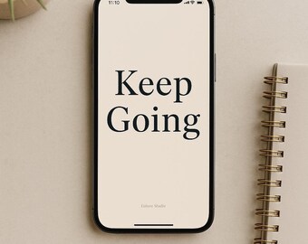 Pode incluir: Um smartphone exibe as palavras "Keep Going" em preto em uma tela creme. Um caderno espiral e uma pequena planta em vaso estão em uma superfície de tons neutros, perto do telefone.