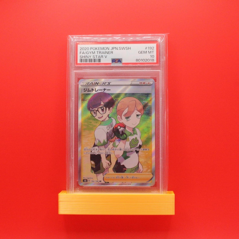 May include: A graded Pok&eacute;mon card with two anime characters, the text "RAIN R'S" and Japanese text. The card is in a clear plastic holder on a yellow stand against a red background. The card is a collectible item.