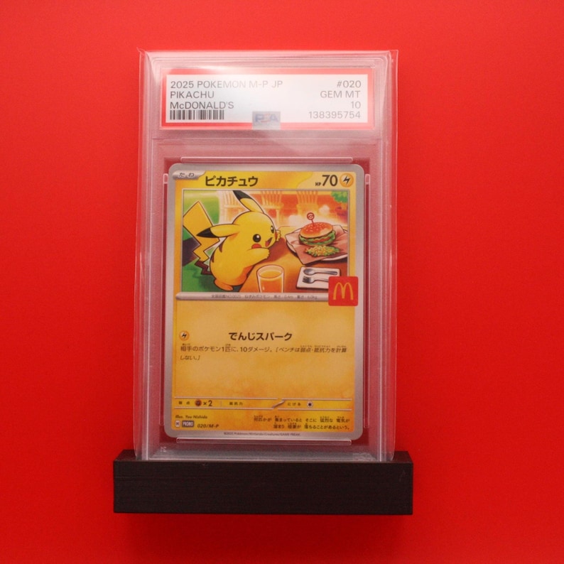 May include: A graded Pok&eacute;mon card featuring Pikachu with a McDonald's theme, encased in a clear holder. The card has Japanese text and a yellow Pikachu illustration. The card is displayed on a black base.