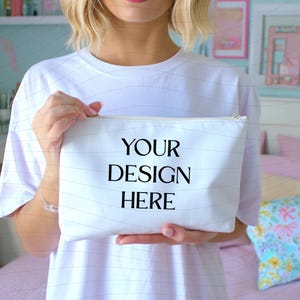 May include: White zippered cosmetic bag with the text "YOUR DESIGN HERE" in black. The bag is held by a person wearing a white t-shirt. The background includes a bed and framed artwork.