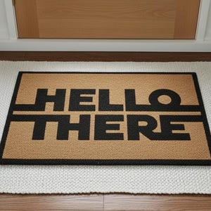 Könnte beinhalten: Eine rechteckige Fußmatte mit den Worten "HELLO THERE" in schwarzen Blockbuchstaben auf beigem Hintergrund. Die Fußmatte liegt auf einem weißen Teppich mit Fransen vor einer Holztür.