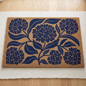 May include: A rectangular doormat with a floral design featuring blue hydrangea blooms and leaves on a tan background. The doormat is placed on a white rug with a fringed edge.