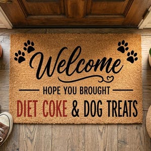 Può includere: Un zerbino in fibra di cocco con la scritta "Welcome" in corsivo, accompagnata da impronte di zampe. Il testo "HOPE YOU BROUGHT DIET COKE & DOG TREATS" è sotto. Scarpe da ginnastica marroni e piante in vaso fiancheggiano il tappeto.