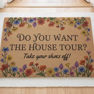 以下が含まれることがあります： 「Do you want the house tour? Take your shoes off!」のテキストと花のボーダーが入った長方形のドアマット。マットはフリンジ付きの白いラグの上にあります。