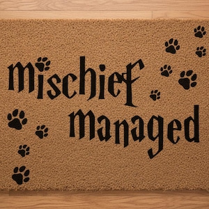 May include: A rectangular doormat with the words "mischief managed" in black gothic font, surrounded by black paw prints on a tan background. The mat is made of a coarse, textured material.