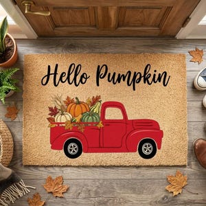 May include: A rectangular tan coir doormat featuring the black text Hello Pumpkin above a graphic of a red pickup truck carrying orange and green pumpkins and autumn leaves. The mat has a coarse, natural fiber texture and a flat, rectangular shape.
