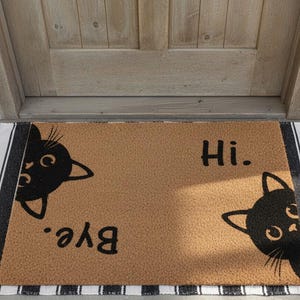 Puede incluir: Una alfombra rectangular con fondo marrón y caras de gato negro a cada lado. Las palabras "Hi." y "Bye." están escritas en negro. La alfombra tiene un borde a rayas blanco y negro.