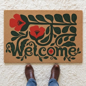 以下が含まれることがあります： 花柄と「Welcome」の文字が入った長方形のドアマット。マットはタン色で、赤い花と緑の葉が描かれています。「Welcome」の文字は緑色の筆記体です。茶色のブーツと青いジーンズが見えます。