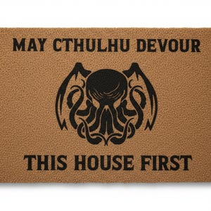 Op de afbeelding: Een rechthoekige deurmat met een bruine achtergrond en zwarte tekst en afbeeldingen. De tekst luidt "MAY CTHULHU DEVOUR THIS HOUSE FIRST." Een zwarte illustratie van een wezen met tentakels en vleugels staat in het midden.