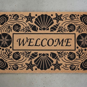 Puede incluir: Una alfombra rectangular con un fondo marrón natural y un diseño de conchas negras. La palabra "WELCOME" está centrada en un marco rectangular. El diseño incluye estrellas de mar, conchas y coral.
