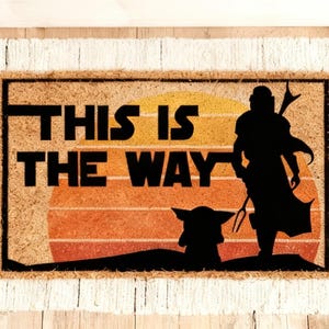 Könnte beinhalten: Rechteckige Fußmatte mit dem Text "THIS IS THE WAY" in Schwarz. Das Design zeigt die Silhouetten einer Figur und einer kleinen Kreatur vor einem Sonnenuntergang. Die Matte hat einen Fransenrand.