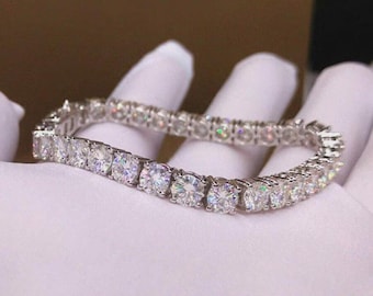 May include: A tennis bracelet with round-cut, clear gemstones set in a silver-colored metal. The bracelet is displayed on a white surface, showcasing the sparkle of the stones. The bracelet is a piece of jewelry.