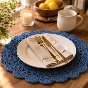 May include: A round, blue crocheted placemat with a scalloped edge, featuring a white ceramic dinner plate, a beige linen napkin, and gold-toned flatware consisting of a fork and a knife placed on top of the plate.