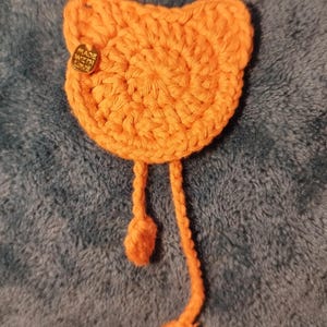 Puede incluir: Un objeto naranja brillante con forma de gato tejido a ganchillo con dos hebras trenzadas colgantes. Una pequeña etiqueta dorada está adherida a la cabeza del gato. El artículo está hecho a mano, posiblemente un accesorio o decoración.