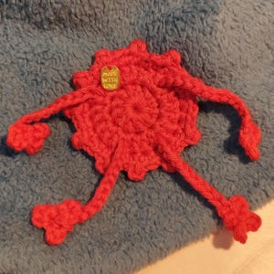May include: A handmade red crochet creature with a circular body and extended arms and legs. A small gold tag with the words "MADE WITH LOVE" is attached to the body. The creature is set against a blue, textured background.