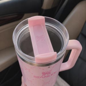 May include: A pink Stanley mug with a handle and a clear lid. The lid has a pink slider to close the opening. The mug has a pink and white pattern and the Stanley logo is visible. The mug is designed to keep beverages hot or cold.