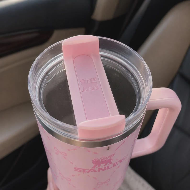 May include: A pink Stanley mug with a handle and a clear lid. The lid has a pink slider to close the opening. The mug has a pink and white pattern and the Stanley logo is visible. The mug is designed to keep beverages hot or cold.