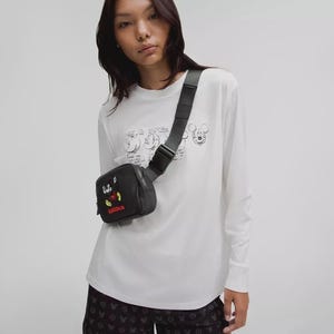 以下が含まれることがあります： ミッキーマウスのデザインの黒いファニーパックが体にかけられています。 パックには赤と黄色の刺繍デザインと「MARATHON」の赤い文字があります。 白い長袖シャツと黒いショートパンツを着ています。 ミッキーマウスのスケッチが描かれています。