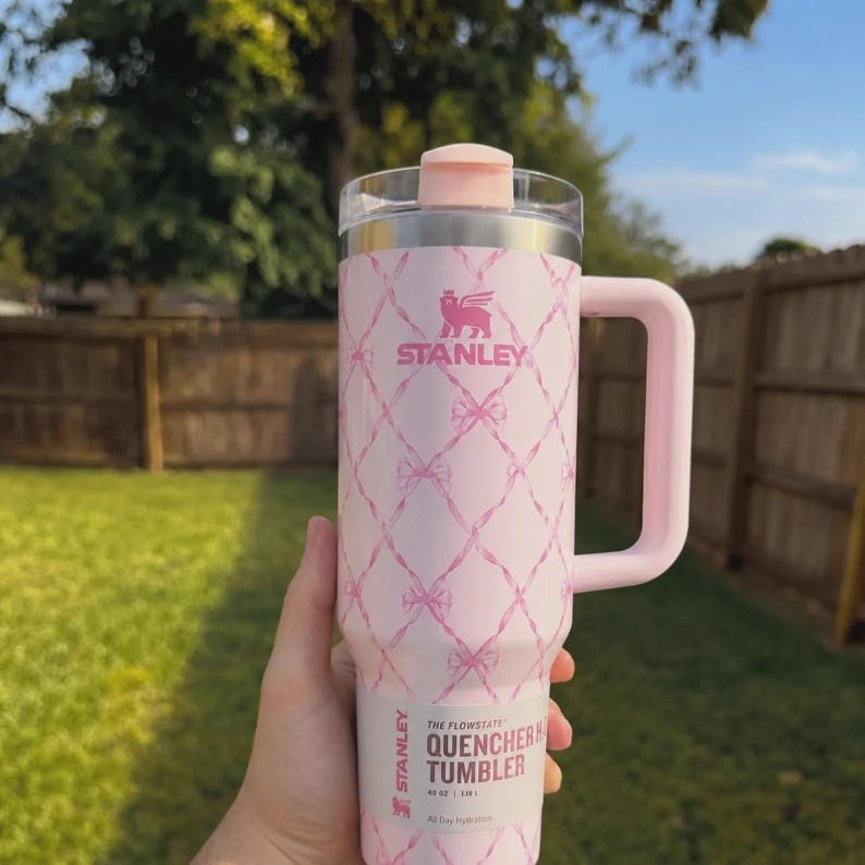 May include: Pink Stanley Quencher tumbler with a handle and a clear lid. The tumbler features a pink bow pattern and the Stanley logo. The text "THE FLOWSTATE QUENCHER TUMBLER" is printed on the bottom.