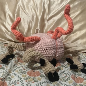 May include: A handmade crochet creature with a pink body, coral-colored tendrils, and tan legs with black feet. The plush toy is displayed on a floral patterned bedspread with a satin pillow in the background. The toy is a unique, whimsical creation.