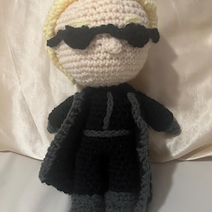 May include: A handmade crochet doll featuring a light skin tone head with blonde hair and black sunglasses. The doll is wearing a black outfit with a gray trim. The doll is approximately 8 inches tall.