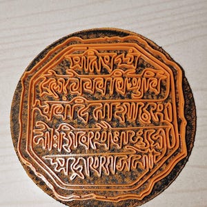 May include: An orange, circular stamp with intricate text in a stylized font. The stamp is set against a dark, textured background. The text is raised and appears to be a religious or cultural inscription.
