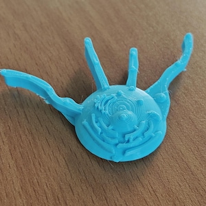 May include: A light blue, 3D-printed object with a circular base and multiple curved, finger-like extensions. The central disc features concentric circles and detailed patterns. The object is resting on a wooden surface.