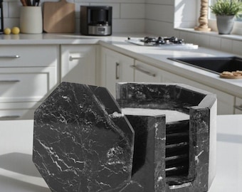 May include: A black marble coaster set with a matching lid. The coasters are octagonal and feature white veining throughout the dark stone. The set is displayed on a white countertop in a kitchen setting.