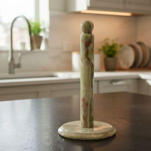 May include: A green and beige marble paper towel holder. The holder has a round base and a tall, cylindrical body with a rounded top. The marble has a natural, swirled pattern. The holder is on a dark countertop in a kitchen setting.