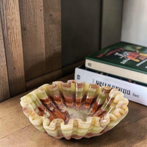 May include: A decorative, ruffled-edge bowl made of onyx. The bowl features a mix of colors, including green, brown, and cream, with a wavy, textured design. The bowl is sitting on a wooden surface.