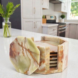May include: A set of hexagonal coasters made of natural stone, featuring shades of green, brown, and cream. The coasters are stacked in a matching holder, showcasing a marbled pattern. The set is displayed on a white countertop.