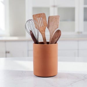 May include: A cylindrical terracotta-colored utensil holder containing a collection of kitchen tools, including a metal wire whisk, a wooden slotted turner, a solid wooden spatula, and two dark brown wooden spoons arranged upright.