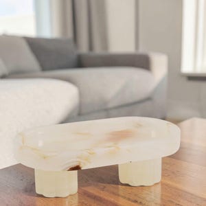 May include: A small, oblong, cream-colored decorative table with three short, rounded legs. The table has a smooth surface with subtle brown and beige veining, and is set on a wooden surface.