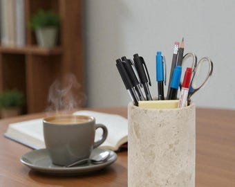 May include: A beige stone pen holder filled with pens, pencils, and scissors sits on a wooden table. A cup of coffee with steam rising from it is on a saucer with a spoon. An open book is in the background.