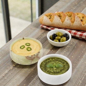 May include: A wooden table displays a culinary arrangement. It features a bowl of yellow dip with green pepper slices, a bowl of green pesto, a bowl of olives, and a loaf of bread. A red and white checkered cloth is under the bread.