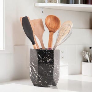 May include: A hexagonal black marble utensil holder with white veining, containing a black silicone spatula, a light brown wooden spatula, a light brown wooden spoon, and a silver metal wire whisk, all standing upright in the container.
