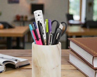 May include: A cylindrical, off-white marble pen holder filled with various pens, pencils, a ruler, and scissors. The holder sits on a wooden surface next to two stacked books with brown covers. The pens include black, blue, pink, green, and purple.