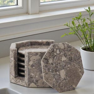 May include: A set of gray marble coasters with a matching holder. The coasters are octagonal and feature a unique pattern of gray and beige veining. The holder is also octagonal and holds several coasters, perfect for a coffee table or bar.