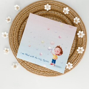 Può includere: Una carta quadrata con un'illustrazione di un bambino che soffia bolle, circondato da bolle dai colori pastello. Il testo "I am filled with the Holy Spirit" è stampato sulla carta. La carta è posizionata su un tappetino intrecciato con accenti floreali bianchi.