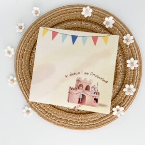 May include: A square greeting card with the text "In Jesus I am Protected" and a pink castle illustration. The card has a banner of colorful flags at the top. The card is on a woven placemat with white flower accents.