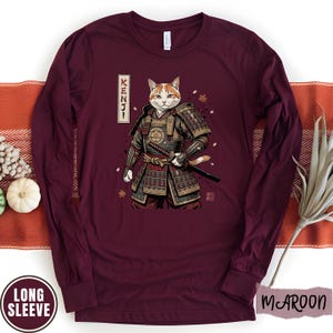 Puede incluir: Camiseta de manga larga color granate con un dise&ntilde;o de gato samur&aacute;i y la palabra japonesa "KENJI". El gato lleva una armadura tradicional y sostiene una espada. La camiseta tiene una etiqueta "LONG SLEEVE".