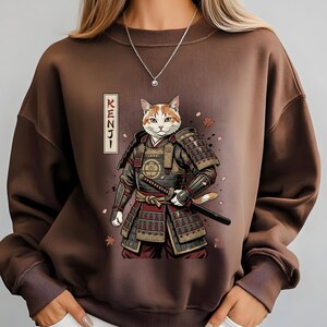 Puede incluir: Sudadera marr&oacute;n con una ilustraci&oacute;n detallada de un gato con armadura de samur&aacute;i, sosteniendo una katana. El gato tiene pelaje naranja y blanco. La palabra "KENJI" est&aacute; impresa verticalmente en el lado izquierdo de la imagen.