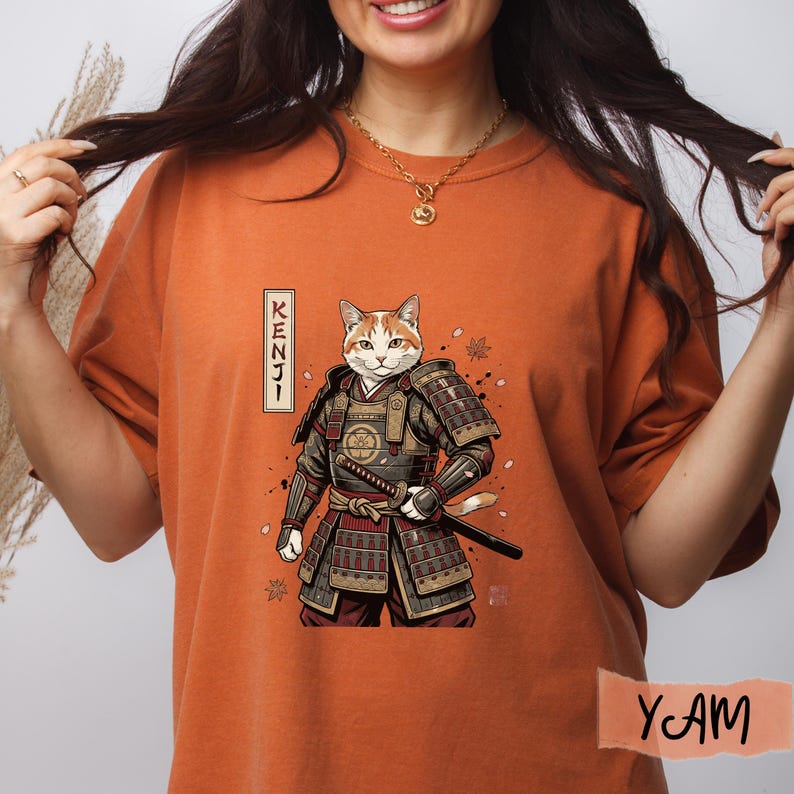 Puede incluir: Camiseta naranja con una ilustraci&oacute;n de un gato guerrero. El gato est&aacute; representado con armadura de samur&aacute;i, sosteniendo una katana. La palabra "KENJI" est&aacute; escrita verticalmente a la izquierda. La camiseta tiene un corte holgado y mangas cortas.