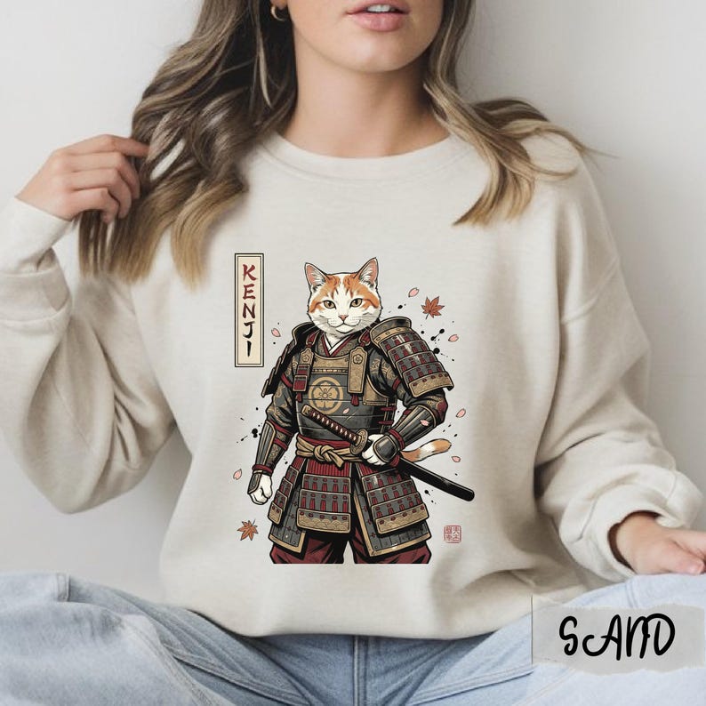 Puede incluir: Sudadera beige con un gato de dibujos animados con armadura de samur&aacute;i, sosteniendo una katana. El gato tiene pelaje naranja y blanco. La palabra "KENJI" est&aacute; en un rect&aacute;ngulo vertical. Tambi&eacute;n hay hojas de oto&ntilde;o y un sello japon&eacute;s.
