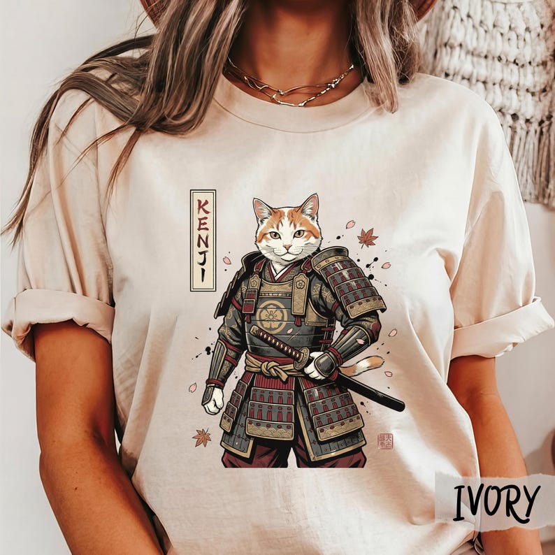 Puede incluir: Camiseta color marfil con una ilustraci&oacute;n de un gato guerrero. El gato est&aacute; representado con una armadura de samur&aacute;i, sosteniendo una katana. La palabra "KENJI" est&aacute; escrita verticalmente en caracteres japoneses. Hojas de oto&ntilde;o dispersas.