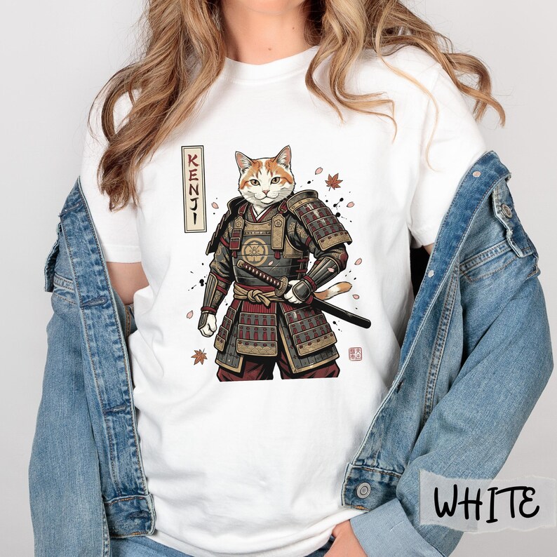 Puede incluir: Camiseta blanca con un gato de dibujos animados con armadura de samur&aacute;i, sosteniendo una katana. El dise&ntilde;o incluye la palabra "KENJI" en caracteres japoneses y hojas de oto&ntilde;o. El gato tiene pelaje naranja y blanco.