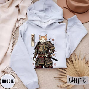 Puede incluir: Sudadera con capucha blanca con una ilustraci&oacute;n detallada de un gato samur&aacute;i, con la palabra "KENJI" en una pancarta vertical. El gato lleva una armadura y sostiene una katana. La palabra "HOODIE" est&aacute; impresa en la parte inferior izquierda. La palabra "WHITE" est&aacute; impresa en la parte inferior derecha.
