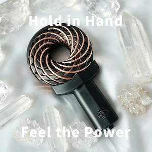 Puede incluir: Un dispositivo de mano negro con una cabeza circular que presenta un diseño de alambre en espiral de color cobre. El mango es negro con un pequeño logotipo en la base. El texto "Hold in Hand" y "Feel the Power" está superpuesto en la imagen.