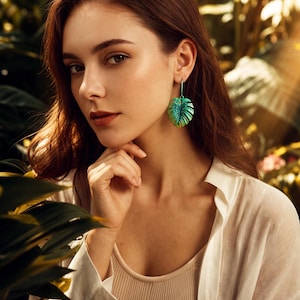 May include: A dangling earring shaped like a monstera leaf, featuring a gradient of vibrant green and yellow tones with a textured surface. The earring is suspended from a thin metal hook, showcasing a detailed vein pattern across the leaf surface.