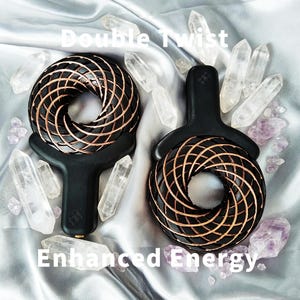 Puede incluir: Dos dispositivos negros de mano con cabezales circulares que presentan un patrón de espiral de alambre de cobre y un mango. El texto Double Twist Enhanced Energy es visible. Los dispositivos están rodeados de cristales de cuarzo y amatista.